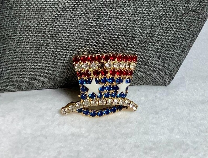 May include: A gold-toned pin with red, white, and blue rhinestones in the shape of a top hat with stars.