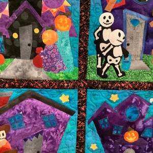 K&ouml;nnte beinhalten: Eine gesteppte Halloween-Wanddekoration mit skurrilen H&auml;usern, K&uuml;rbissen und Skelettfiguren. Die H&auml;user sind lila und schwarz mit verschiedenen Details. Der Hintergrund ist hellblau mit Stern- und Bl&auml;ttermustern.