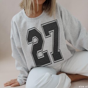 May include: A gray sweatshirt with a black and white number 27 printed on the front. The sweatshirt is a relaxed fit and has long sleeves.