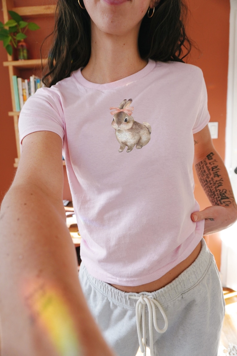 model wears a fitted crop top baby tee with a bunny rabbit on it wearing a pink ribbon bow in the style of coquette cottagecore downtown girl clean girl aesthetic