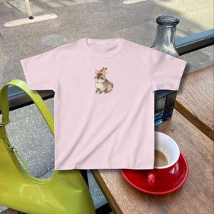 model wears a fitted crop top baby tee with a bunny rabbit on it wearing a pink ribbon bow in the style of coquette cottagecore downtown girl clean girl aesthetic