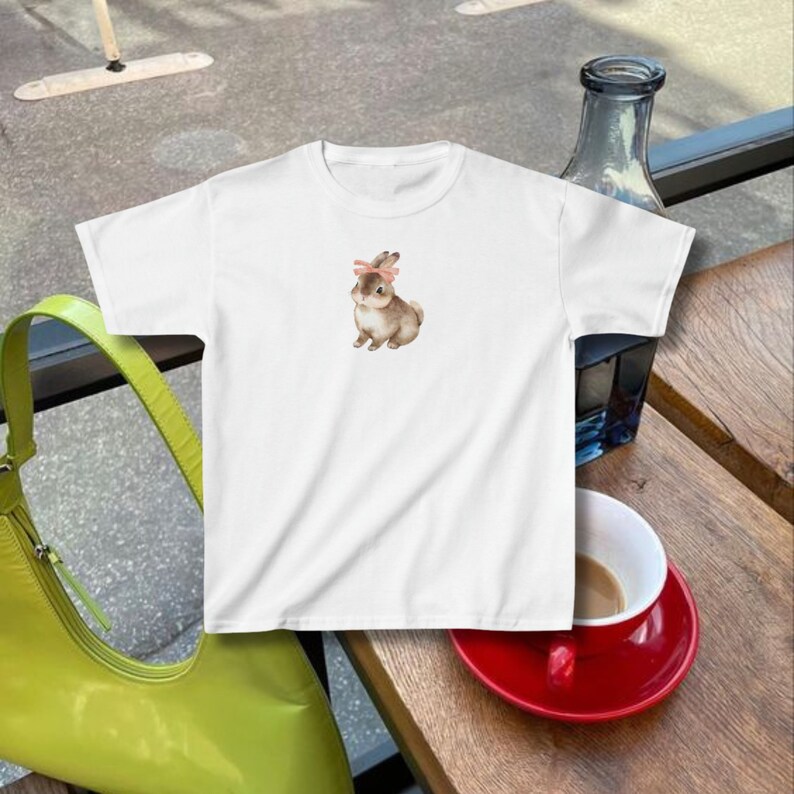 model wears a fitted crop top baby tee with a bunny rabbit on it wearing a pink ribbon bow in the style of coquette cottagecore downtown girl clean girl aesthetic