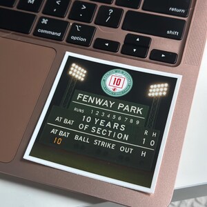 May include: A sticker featuring a Fenway Park scoreboard design, with the text "FENWAY PARK" and "10 YEARS OF SECTION." The sticker is placed on a rose gold laptop, with the keyboard visible.