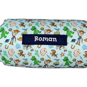 May include: A light blue rolled blanket featuring a repeating pattern of Toy Story characters including Woody, Buzz Lightyear, Rex, and Hamm, along with cacti and stars. A dark blue rectangular patch is centered with the name Roman embroidered in white.
