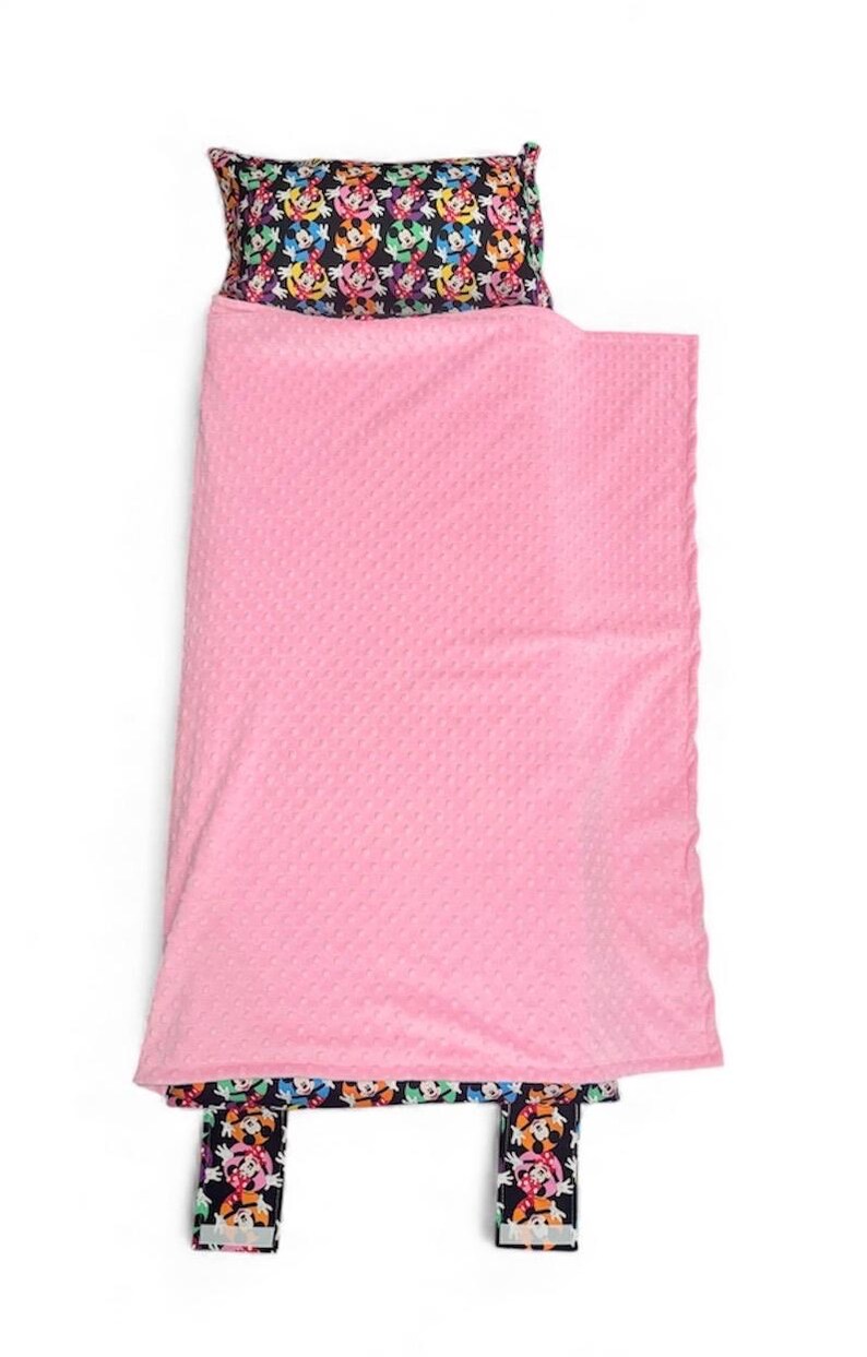 May include: A pink nap mat set with a patterned pillow and footrest. The pillow and footrest feature a colorful repeating pattern of cartoon characters on a black background. The blanket is a solid pink color.