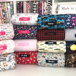 May include: A display of personalized bolster pillows with various fabric patterns, including unicorns, dinosaurs, and trucks. Each pillow has a name tag, such as "Decker" and "Annalynn". The background shows shelves of fabric rolls.