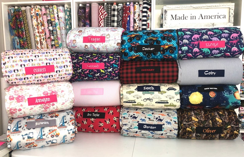 May include: A display of personalized bolster pillows with various fabric patterns, including unicorns, dinosaurs, and trucks. Each pillow has a name tag, such as "Decker" and "Annalynn". The background shows shelves of fabric rolls.