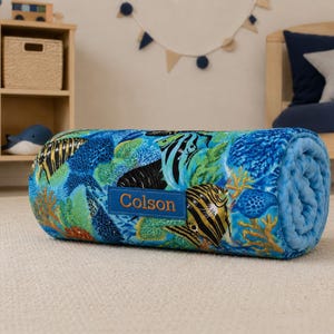 May include: A rolled-up plush blanket featuring a vibrant underwater scene with colorful tropical fish, coral reefs, and sea plants in shades of blue, green, and orange. A rectangular blue fabric patch is sewn on the front with the name Colson.