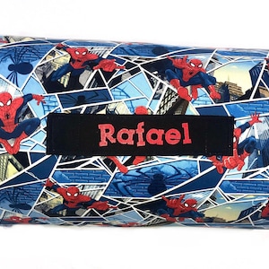 May include: A cylindrical bolster pillow featuring a blue and white comic book print of Spider-Man in various action poses. A rectangular black fabric patch is centered on the pillow with the name Rafael embroidered in red thread.