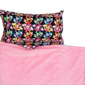 May include: A black pillowcase with a repeating pattern of Mickey and Minnie Mouse in colorful circles. The pillow rests on a pink, textured blanket. The blanket is a solid color and has a soft, raised dot pattern.