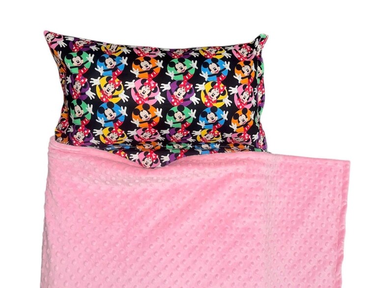 May include: A black pillowcase with a repeating pattern of Mickey and Minnie Mouse in colorful circles. The pillow rests on a pink, textured blanket. The blanket is a solid color and has a soft, raised dot pattern.