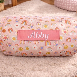 May include: A cylindrical bolster pillow in a soft pink hue, featuring a repeating pattern of colorful rainbows, suns, and daisies. A rectangular pink fabric patch is centered on the front, displaying the name Abby in white cursive lettering.
