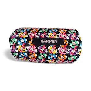 May include: A black, cylindrical pillow with a colorful pattern of Minnie Mouse characters. The pillow has a black label with the name "HARPER" in white letters. The design features Minnie Mouse in various colors, including red, yellow, blue, and purple.