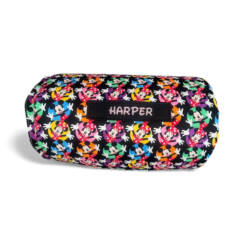 May include: A black, cylindrical pillow with a colorful pattern of Minnie Mouse characters. The pillow has a black label with the name "HARPER" in white letters. The design features Minnie Mouse in various colors, including red, yellow, blue, and purple.