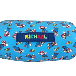 May include: A cylindrical bolster pillow featuring a light blue fabric with a repeating pattern of Super Mario characters and logos. A dark blue rectangular fabric patch is centered on the front with the name MICHAEL printed in multicolored letters.
