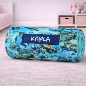 May include: A rolled-up blanket featuring a vibrant blue and green sea turtle pattern. A dark blue rectangular patch is centered on the front, displaying the name KAYLA in white capital letters. The blanket has a soft, plush texture throughout.