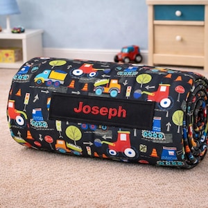 May include: A rolled-up black fabric mat featuring a colorful construction vehicle pattern with tractors, dump trucks, and traffic cones. A rectangular black patch is centered on the front with the name Joseph embroidered in red thread.