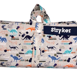 May include: A gray and white fabric play mat with a dinosaur print. The mat folds up and has a handle. The mat has a blue name tag with the name "Stryker" printed in white.