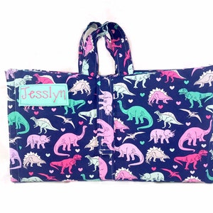 May include: A blue canvas tote bag with a pink and green dinosaur print and hearts. The bag has a personalized name tag that says "Jesslyn".