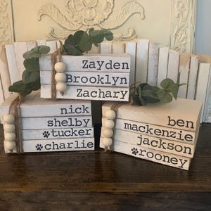 May include: Three stacked wooden blocks with names printed in black. The blocks are white with a distressed finish, and are decorated with wooden beads and faux greenery. Names include: Zayden, Brooklyn, Zachary, Nick, Shelby, Tucker, Charlie, Ben, Mackenzie, Jackson, and Rooney.