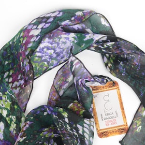 Op de afbeelding: Een doorschijnende sjaal met een bloemenpatroon in groen, paars en wit. De sjaal is gedrapeerd op een witte ondergrond, met een label met de tekst "ERICA WADDELL". De sjaal is gemaakt van een lichtgewicht materiaal.