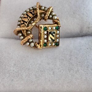 May include: A gold-toned brooch with a circular design, featuring the letters "AIKY" in black. The brooch is adorned with small pearls and a square element with the letter "Z". Green and white stones are set around the square.