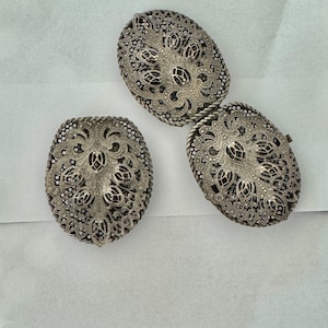 May include: Two silver filigree oval-shaped belt buckles with intricate floral designs.
