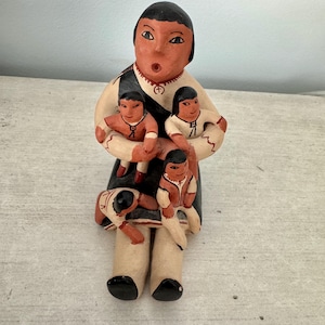 May include: Hand-painted ceramic figurine of a mother with four children. The mother is sitting with her legs crossed and holding two children in her arms. Two more children are sitting on her lap. The figurine is painted in shades of brown, black, white, and red.