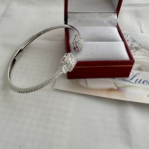 May include: A silver bangle bracelet with a pave diamond setting and two square diamond accents. The bracelet is in a red box.