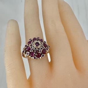 May include: A gold-toned ring featuring a cluster of deep red gemstones. The ring's design showcases a central, larger stone surrounded by smaller, similarly colored gems, creating a floral or cluster arrangement. The ring is set on a light-toned background.