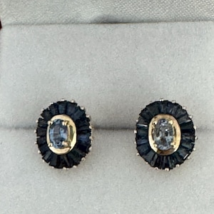 May include: A pair of oval-shaped stud earrings. Each earring features a light blue gemstone set in a gold-colored frame, surrounded by a dark blue, spiky border. The earrings are displayed on a light gray surface.