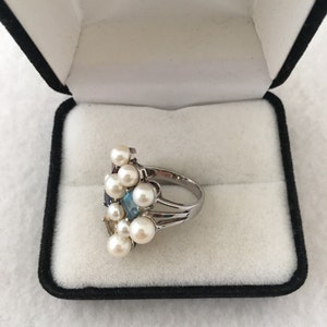 May include: A silver ring featuring a cluster of white pearls and colourful gemstones. The ring is displayed in a black velvet box with the inscription "America's Leading Jeweler" inside.