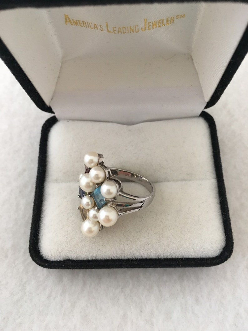 May include: A silver ring featuring a cluster of white pearls and colourful gemstones. The ring is displayed in a black velvet box with the inscription "America's Leading Jeweler" inside.