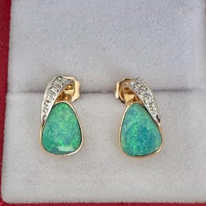 May include: A pair of gold stud earrings with pear-shaped, green opals. The earrings have a gold frame and a curved, diamond-studded accent. The earrings are displayed in a red jewelry box.