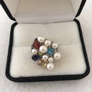 May include: A multi-coloured gemstone and pearl ring in a black velvet ring box. The ring features a cluster of small, square-shaped gemstones in various colours, including red, green, blue, and purple, surrounded by white pearls.