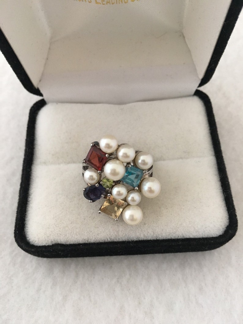 May include: A multi-coloured gemstone and pearl ring in a black velvet ring box. The ring features a cluster of small, square-shaped gemstones in various colours, including red, green, blue, and purple, surrounded by white pearls.