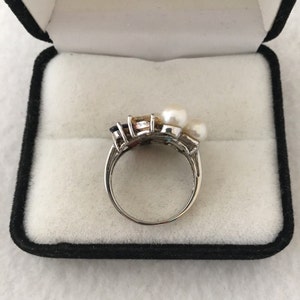 May include: A silver ring featuring a cluster of colourful gemstones and three white pearls. The ring is displayed in a black velvet ring box.