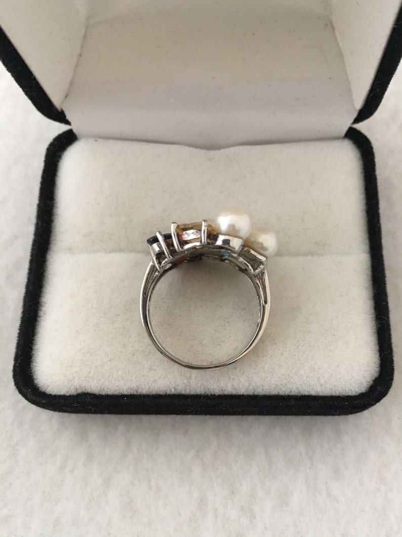 May include: A silver ring featuring a cluster of colourful gemstones and three white pearls. The ring is displayed in a black velvet ring box.