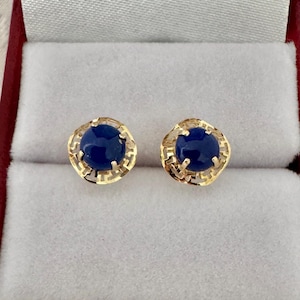 May include: A pair of stud earrings featuring round, deep blue cabochon stones set in gold metal. Each stone is surrounded by a decorative, openwork gold frame with a geometric pattern, secured by four prongs holding the central blue gemstone.