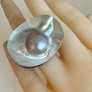 May include: Oval-shaped silver ring with a large, iridescent pearl-like center stone. The ring's setting has a textured, shell-like appearance. The ring is displayed on a hand, showcasing its size and design.