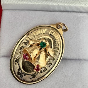May include: An oval gold pendant featuring a raised design of two bells tied with a ribbon. The bells are accented with one small green gemstone and two small red gemstones. The border is engraved with the text MERRY CHRISTMAS.