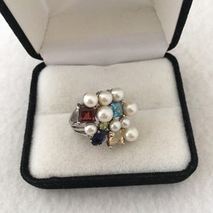May include: A silver ring featuring a cluster of pearls and colourful gemstones, including red, blue, green, and yellow. The ring is presented in a black velvet box with the inscription "America's Leading Jeweler" inside.
