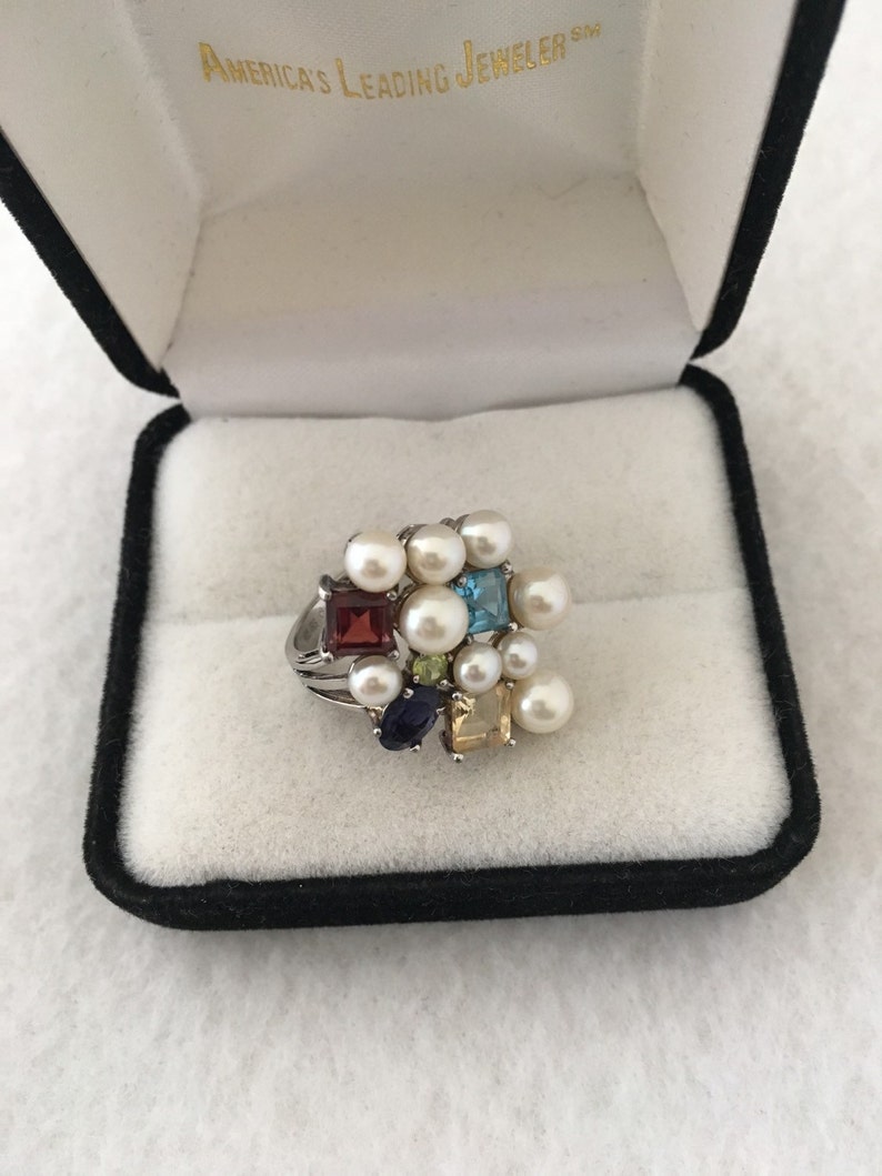 May include: A silver ring featuring a cluster of pearls and colourful gemstones, including red, blue, green, and yellow. The ring is presented in a black velvet box with the inscription "America's Leading Jeweler" inside.