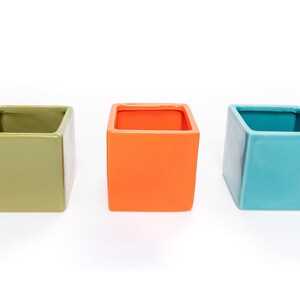 May include: Three square ceramic planters in different colors: green, orange, and blue. The planters are empty and ready to be filled with plants.