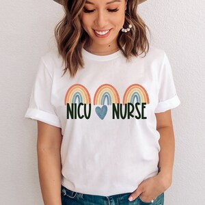 Pu&ograve; includere: Maglietta bianca con tre disegni arcobaleno e il testo "NICU HEART NURSE".