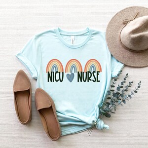 Pu&ograve; includere: Una maglietta blu chiaro con tre disegni di arcobaleno e il testo "NICU HEART NURSE".