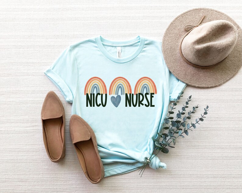 Pu&ograve; includere: Una maglietta blu chiaro con tre disegni di arcobaleno e il testo "NICU HEART NURSE".