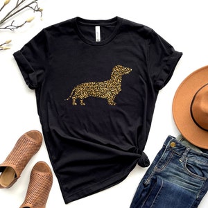 Może przedstawiać: Czarny t-shirt z złotą sylwetką  dackel  zrobioną z nadruku lamparta. Koszulka jest pokazana z brązowym kapeluszem, niebieskimi dżinsami i brązowymi sandałami.
