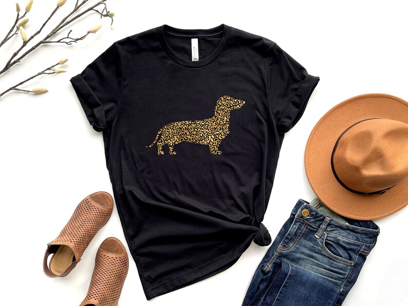Może przedstawiać: Czarny t-shirt z złotą sylwetką  dackel  zrobioną z nadruku lamparta. Koszulka jest pokazana z brązowym kapeluszem, niebieskimi dżinsami i brązowymi sandałami.