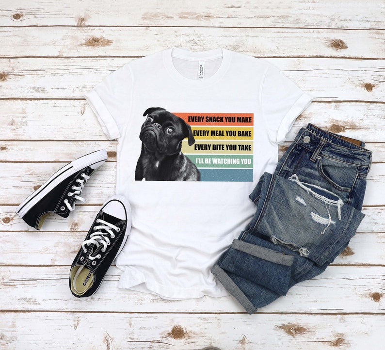 May include: White t-shirt with a black pug dog graphic and the text "EVERY SNACK YOU MAKE EVERY MEAL YOU BAKE EVERY BITE YOU TAKE I'LL BE WATCHING YOU" in a retro rainbow design.